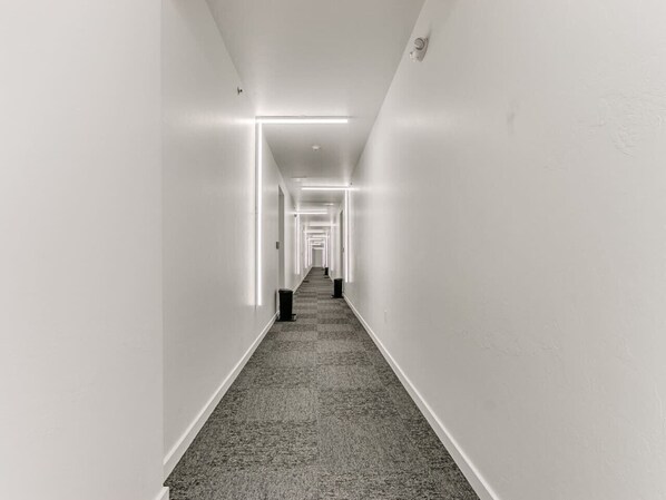 A long, narrow hallway with white walls and a carpeted floor, leading to a door at the end.
