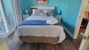 A bedroom with a large bed, a headboard with a beach scene, two bedside tables with lamps, and a view of the outdoors through a sliding door.
