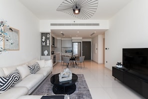 A modern living room with a sofa, coffee table, and TV. There is a dining area with chairs and a table, and a kitchen in the background.