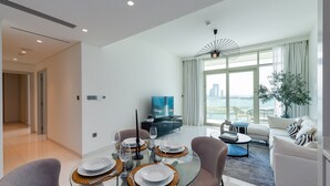 Uma sala de estar moderna com uma mesa de jantar de vidro posta para quatro pessoas, um sofá, uma mesa de centro e uma televisão ao fundo.