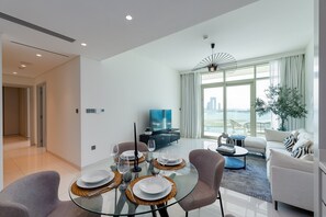 A modern living room with a glass dining table set for four, a sofa, a coffee table, and a TV in the background.