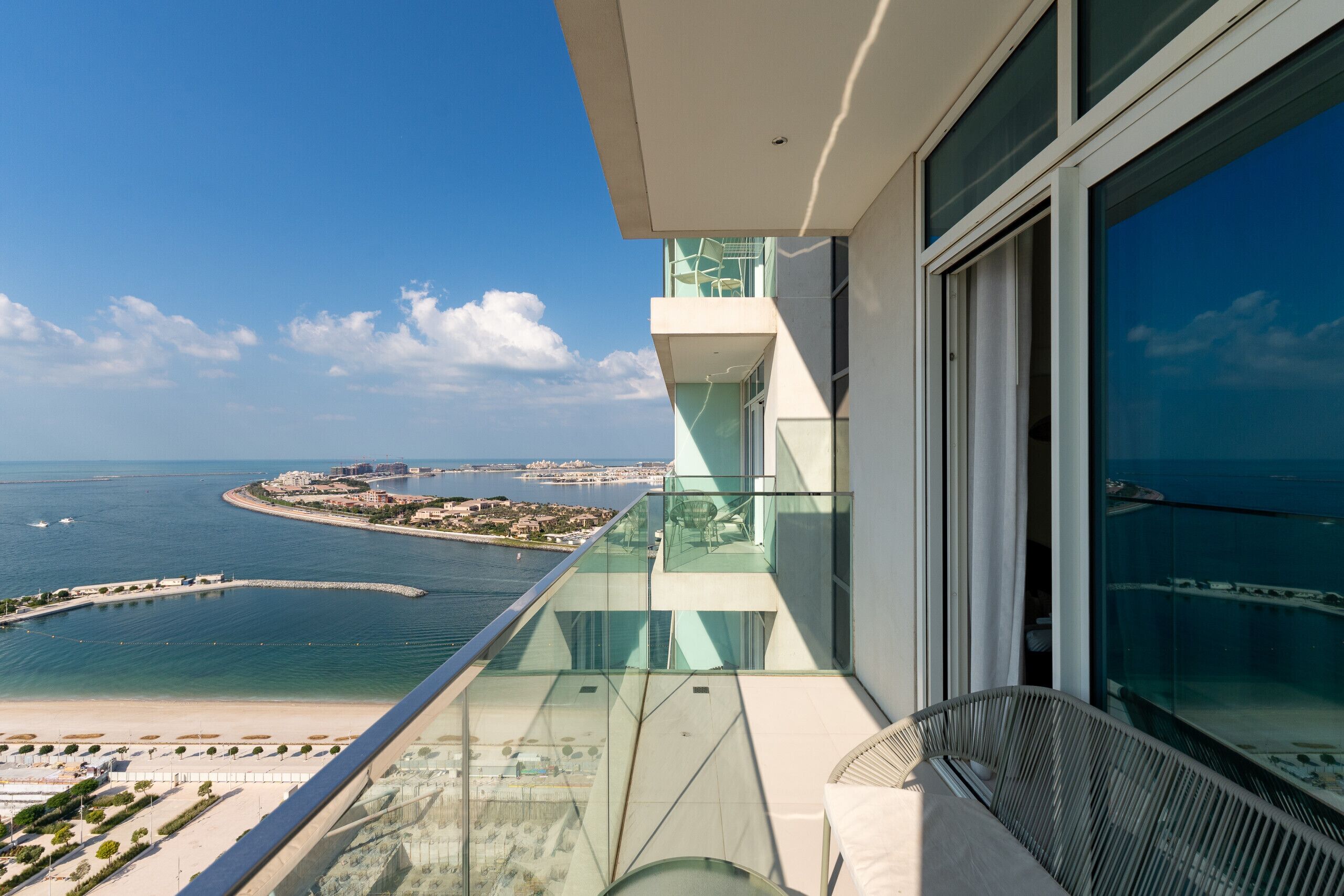 A high-rise building with a glass balcony overlooking a coastal area, including a beach and marina.