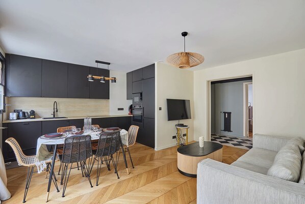 A modern kitchen with dark cabinets, a dining table with chairs, and a living area with a sofa and a TV. - Superb Apartement Tour Eiffel Invalides (Paris)