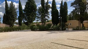 A gravel parking area with tall cypress trees and a yellow building in the background. - Cottage Saint-remy-de-provence (St.-Rémy-de-Provence)