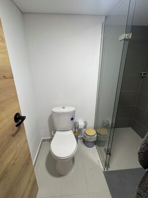 A small, compact bathroom with a toilet, a glass shower enclosure, and a wooden door. - Duplex loft base  (Medellín)