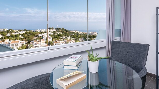 A balcony with a glass table, a potted plant, and books. A view of a coastal town is visible through the window. - Cosy Apartment in Mijas (Mijas)