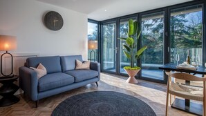 A modern living room with a sofa, a round rug, a dining table with chairs, a potted plant, and large windows offering a view of trees. - Wisemans House Apartment 1 - Wisemans Bridge (Saundersfoot)