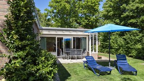 A covered patio with a table and chairs, two sun loungers, and a blue umbrella. - Bungalow in North Holland by the Coast (Sint Maartensvlotbrug)