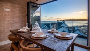Una mesa de comedor dispuesta con platos, vasos y una botella de vino, con vistas a un paisaje marino.