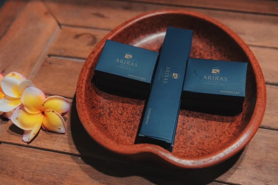 A terracotta bowl containing two blue boxes labeled "ARIRAS" with "SHOWER CAFE" and "SANITARY BAG" on them, placed on a wooden surface with a yellow and white flower beside the bowl.
