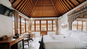 A hotel room with a wooden ceiling, a bed with white linens, a desk with a coffee maker, and a view of the outside through large windows. - The Ariras Resort (Karangasem)