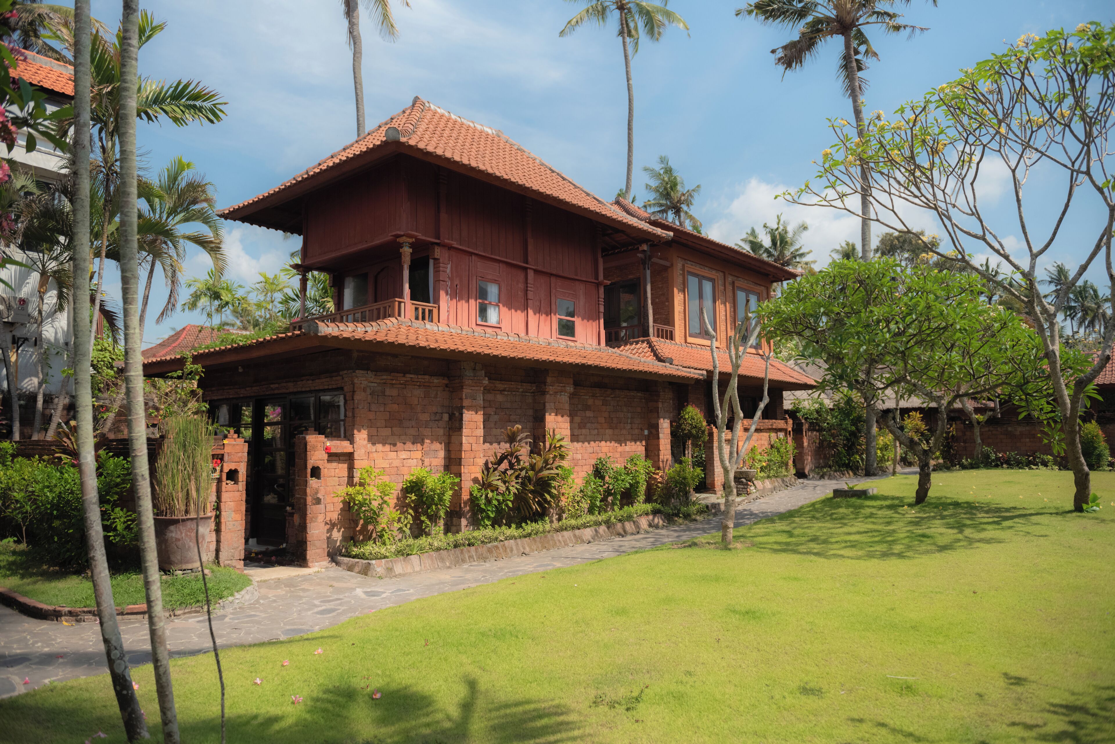 A two-story brick house with a tiled roof, surrounded by a well-maintained lawn and palm trees.