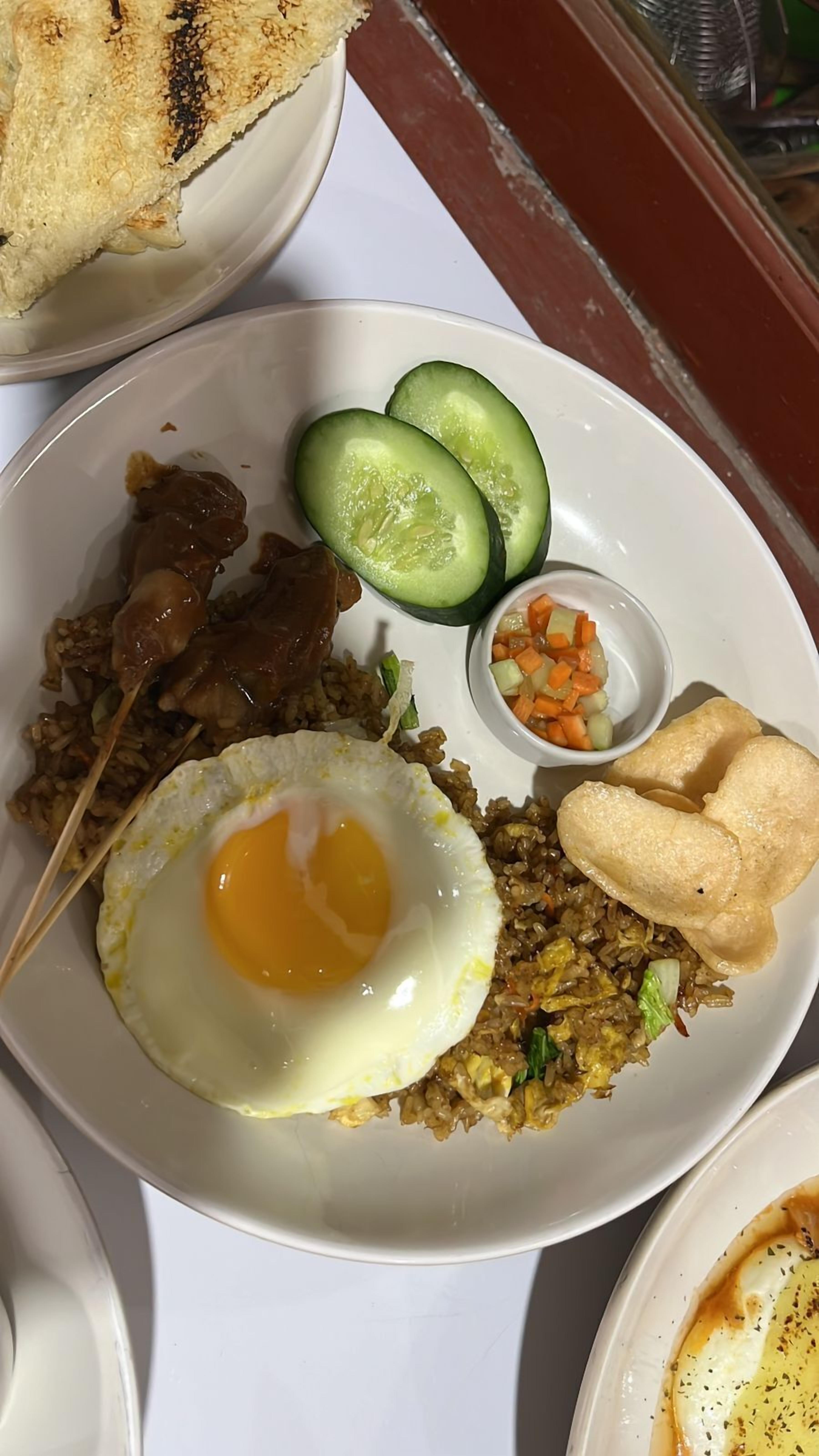A plate of fried rice with a fried egg, cucumber slices, meat skewers, and a small bowl of vegetables.