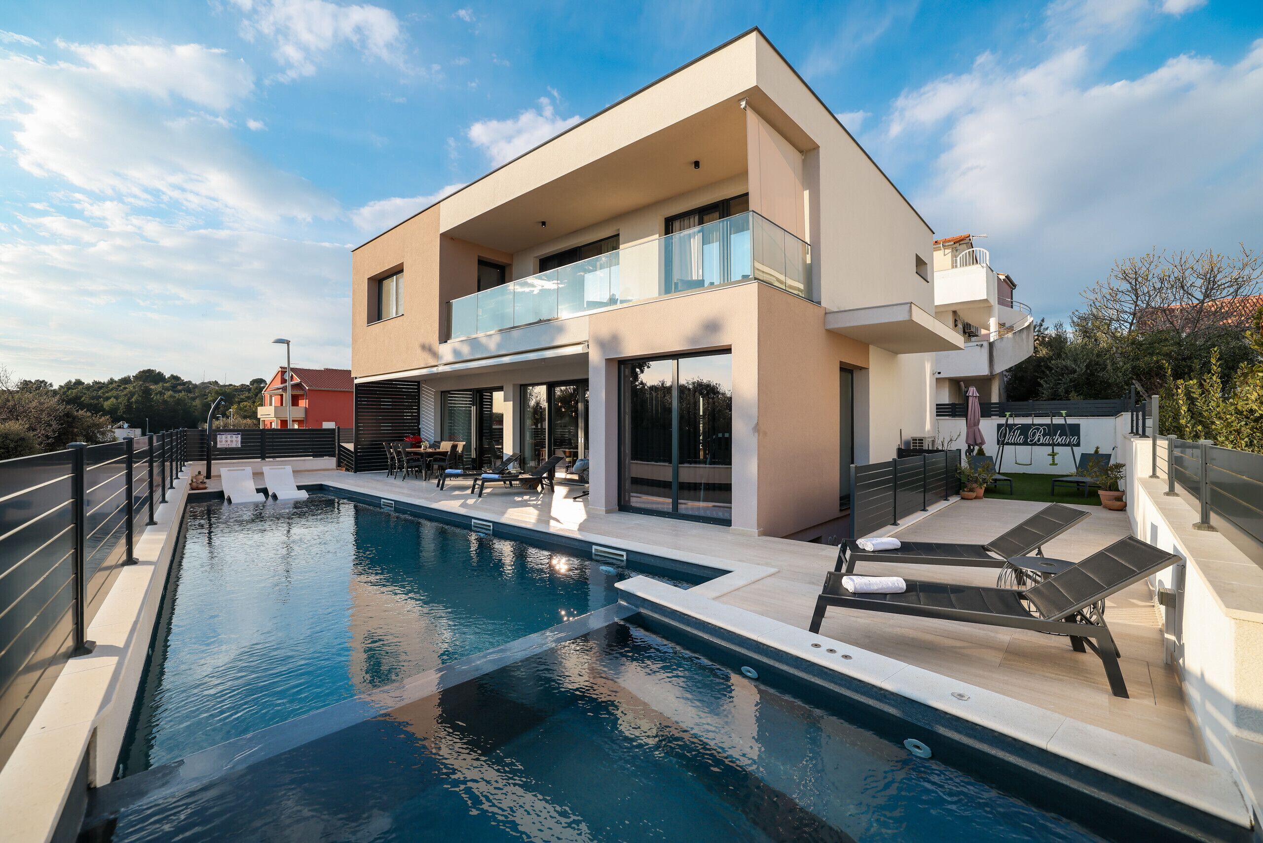 Et moderne hus med to etager, en swimmingpool, udendørs møbler og en balkon.
