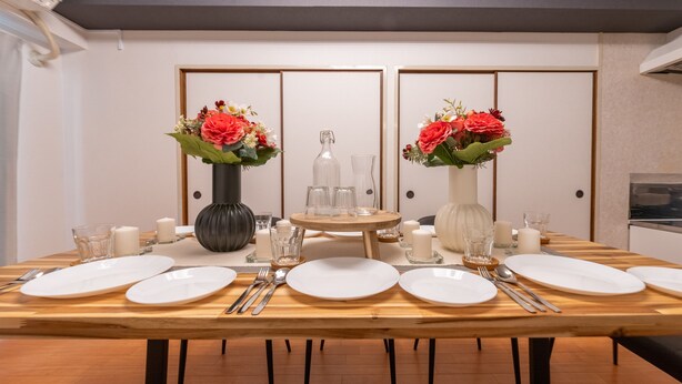 Tavola imbandita per sei persone, con piatti bianchi, posateria e due vasi con fiori rossi.