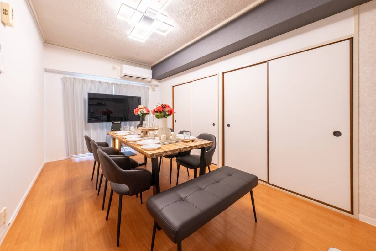 Un comedor con una mesa de madera preparada para cuatro personas, sillas negras y un banco. Hay un jarrón con flores sobre la mesa.