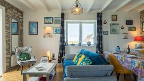 A cozy living room with a sofa, armchairs, a coffee table, and a dining table set for two. There is a large window with curtains, a stone wall, and various framed pictures on the walls. - La Grande Roche by Interhome (Porspoder)