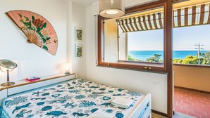Quarto com cama, janela com vista para o mar, um ventilador pendurado e uma luminária.