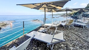 Uma área de descanso à beira-mar com espreguiçadeiras e guarda-sóis, com vista para o mar e a costa rochosa.