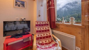 A cozy room with a television, a red entertainment unit, a patterned chair, a window with a view of mountains, and a framed picture on the wall.