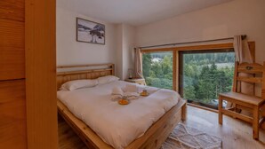 A bedroom with a wooden bed, a nightstand, a chair, a large window with a view of trees, and a framed picture on the wall.