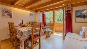 A wooden interior with a dining table, chairs, a sofa, and a window with a view of trees.