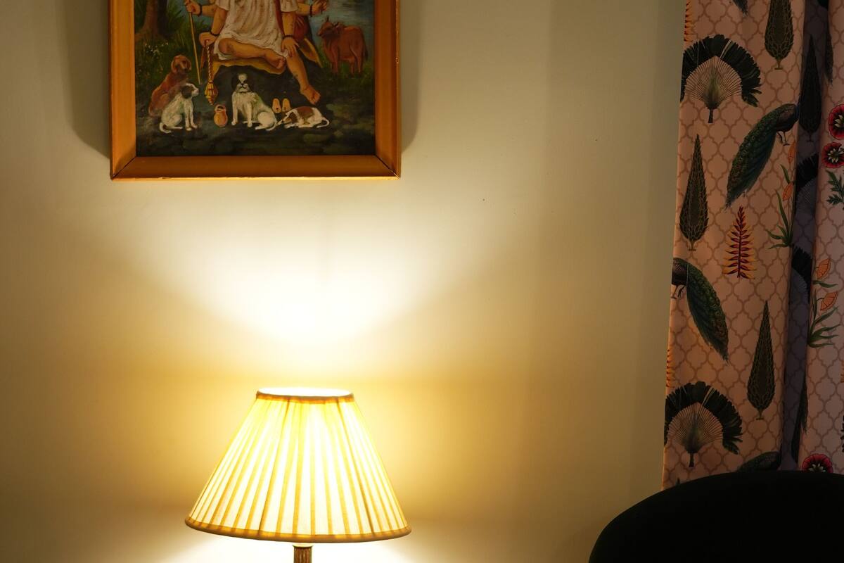 Una mesa con una lámpara, un cuadro enmarco y una cortina con estampado de hojas.