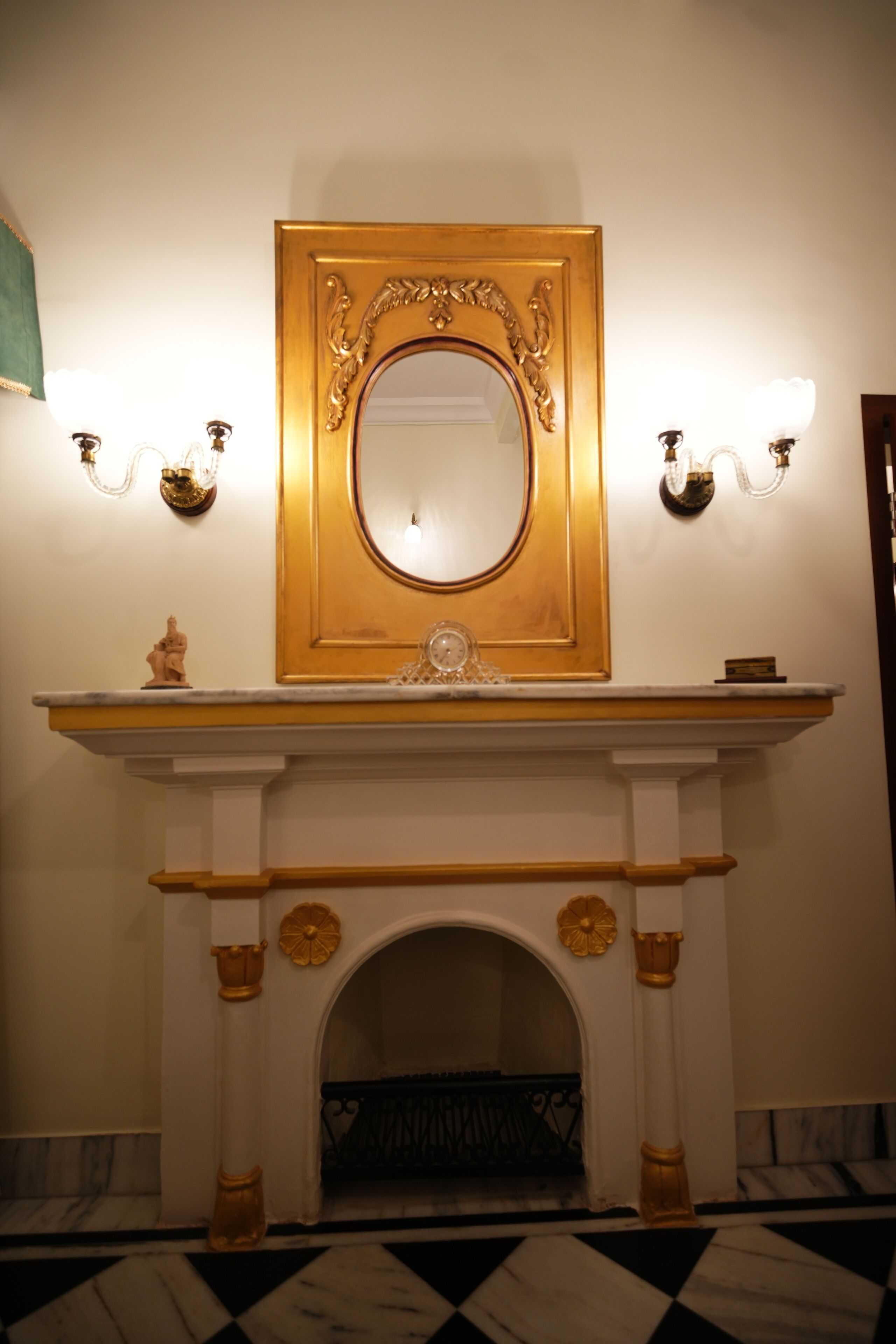 A grand hallway with a marble floor, a white fireplace with gold accents, and a large ornate mirror above it.