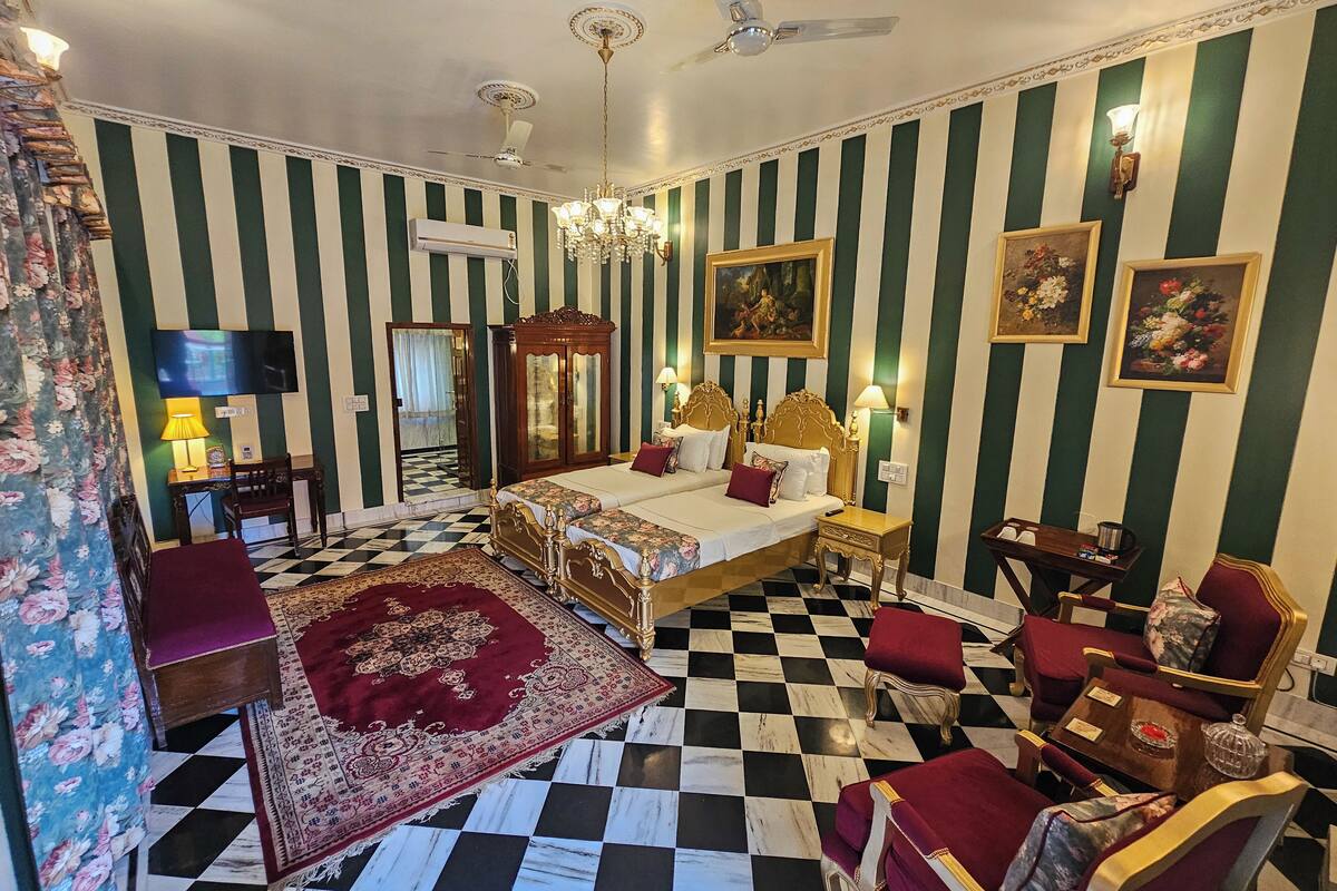 Una habitación de hotel con piso a cuadros, dos camas con cabeceros ornamentados, un candelabro y un espejo grande.