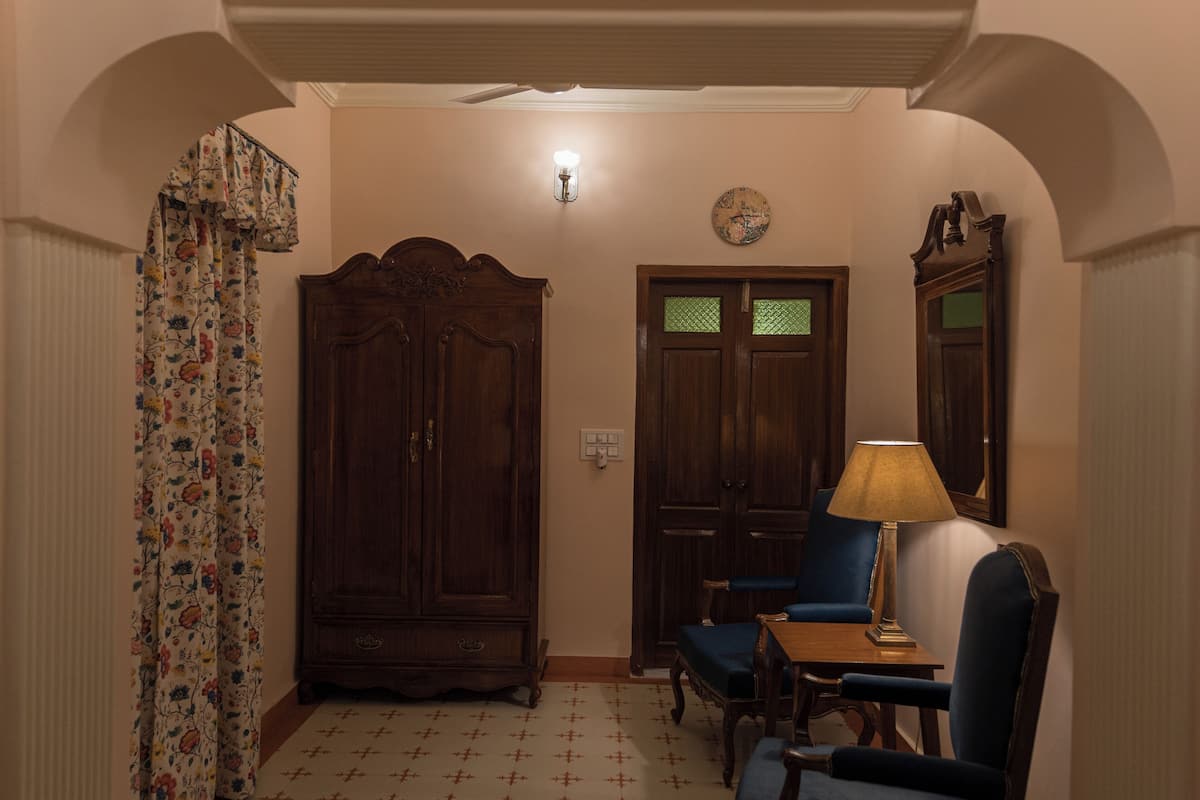 Una habitación con un armario de madera, un sillón azul, una mesa con lámpara y una cortina con motivos florales.
