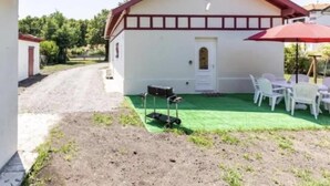 En enplansbyggnad med en vit fasad och ett rött tak. Det finns en liten veranda med en dörr och ett fönster, samt en liten yta med konstgräs och utemöbler.