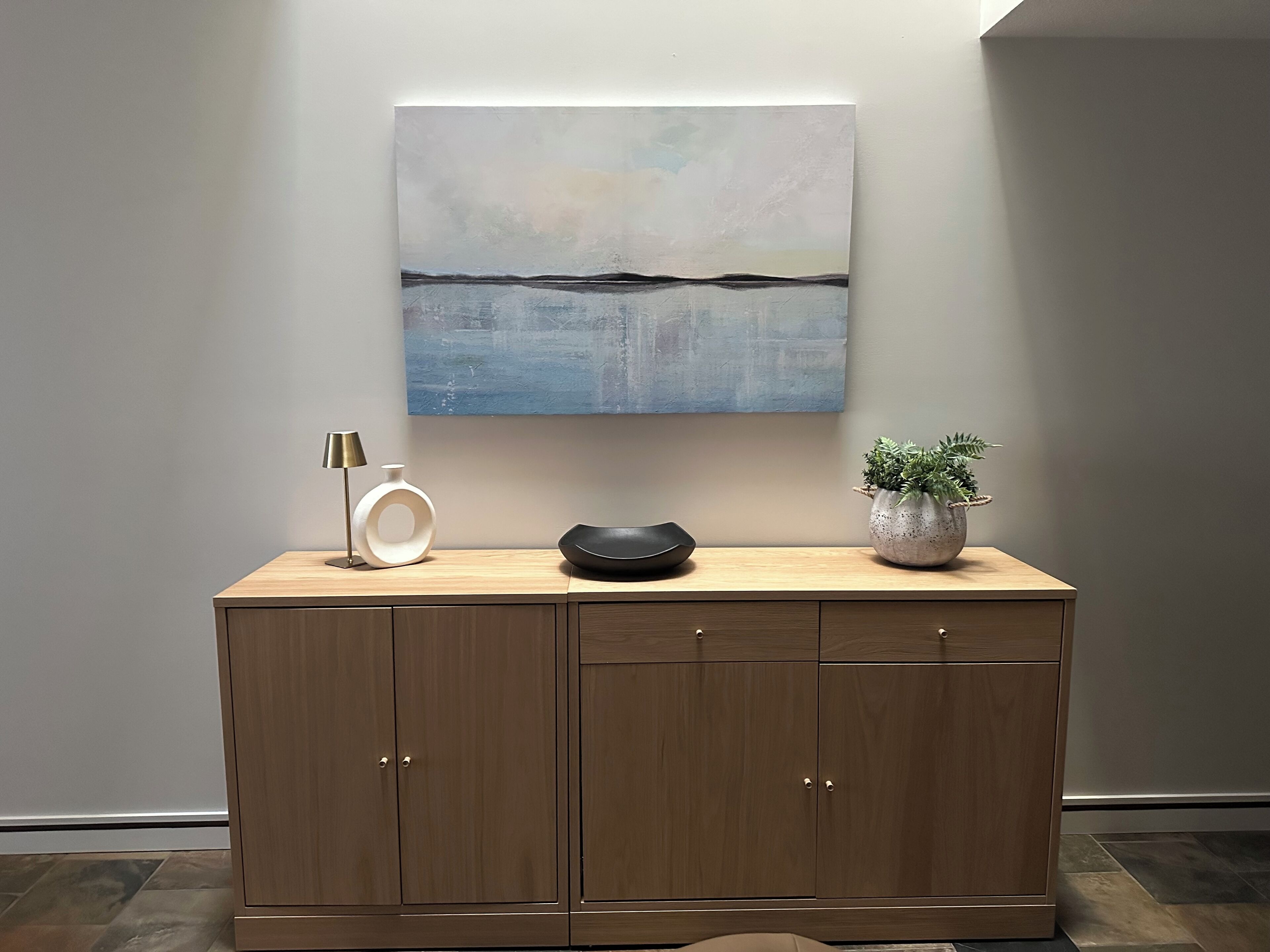 A wooden cabinet with a minimalist design, a white vase, a black bowl, a small potted plant, and a modern abstract painting on the wall above.
