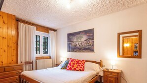 A wooden bed with a headboard, a bedside table, a nightstand, a window with curtains, and a wall-mounted painting. - Casa Rossa by Interhome (Madesimo)