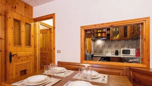 A dining room with a wooden table set for four, a kitchen visible through a window, and wooden paneling on the walls. - Casa Rossa by Interhome (Madesimo)