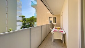 A narrow balcony with a white table and chairs, a small window, and a view of other buildings and trees.