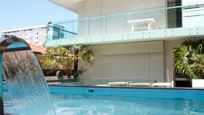 A swimming pool with a water feature, surrounded by a modern building with glass railings and palm trees.