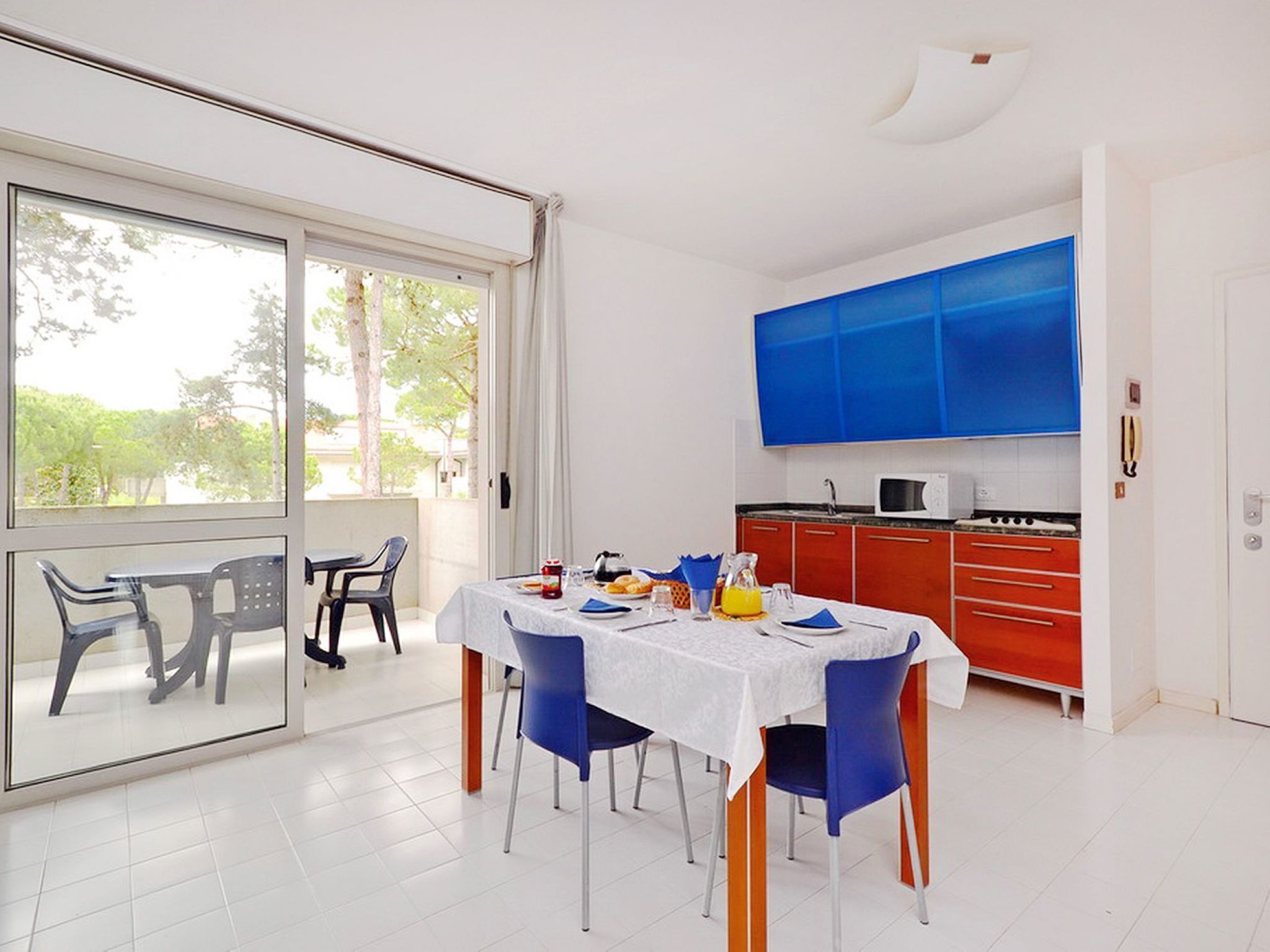 A dining area with a table set for two, a kitchen with red cabinets, and a large window with a view of trees.