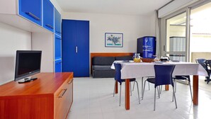A modern room with a wooden desk, a television, a dining table with chairs, a blue cabinet, and a large window. - Parco Hemingway by Interhome (Lignano Pineta)