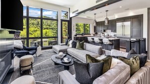 A modern kitchen with stainless steel appliances, a dining area with a bar, and a living room with a large window overlooking greenery.