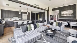 Uma cozinha moderna com armários escuros, uma sala de jantar com mesa e cadeiras brancas, uma sala de estar com sofá cinza e tapete estampado, e um quadro abstrato grande na parede.