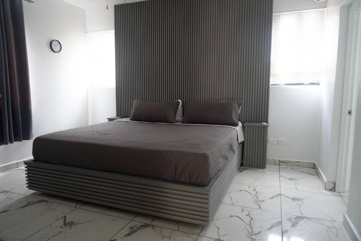 Un dormitorio moderno con una cama grande, una mesita de noche, un reloj en la pared y una ventana con persianas.