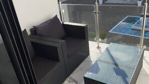 Egy erkély, melyen fekete, fonott műanyagból készült kanapé, egy kis asztal és üvegkorlát található.