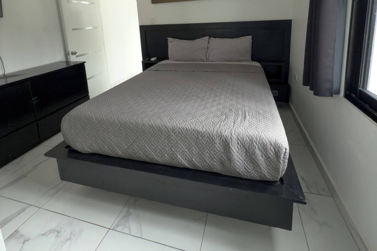 Un dormitorio moderno con una cama, un ventilador de techo, un cuadro de una puesta de sol, un armario y una ventana con cortinas.