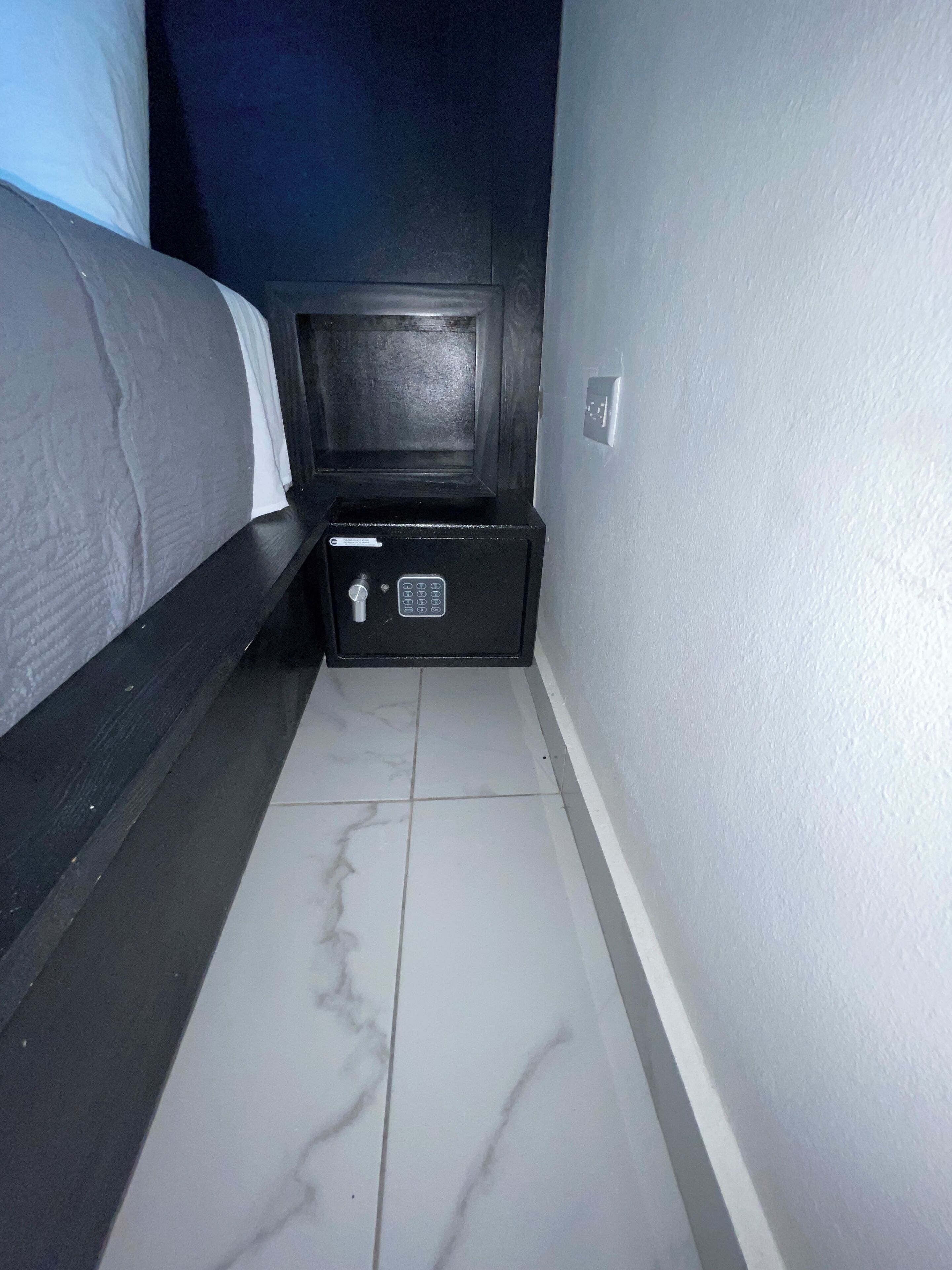 A narrow hallway with a bed on the left, a small black safe, and a white tiled floor.