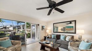 A spacious living room with a large ceiling fan, a glass sliding door leading to a balcony with a view of palm trees and a building, and a large mirror on the wall.