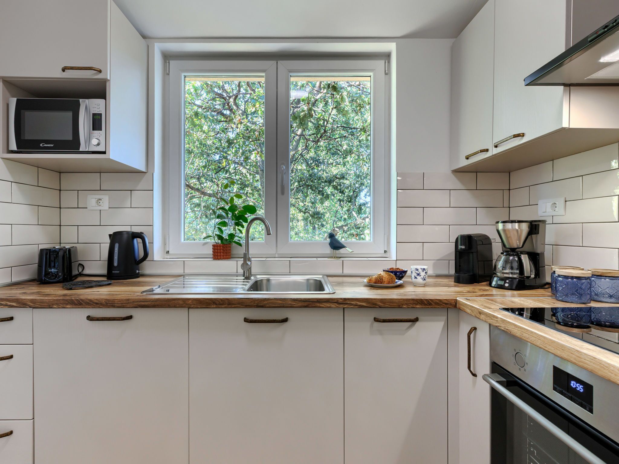 A modern kitchen with a microwave, toaster, kettle, coffee maker, and a window with a view of trees.