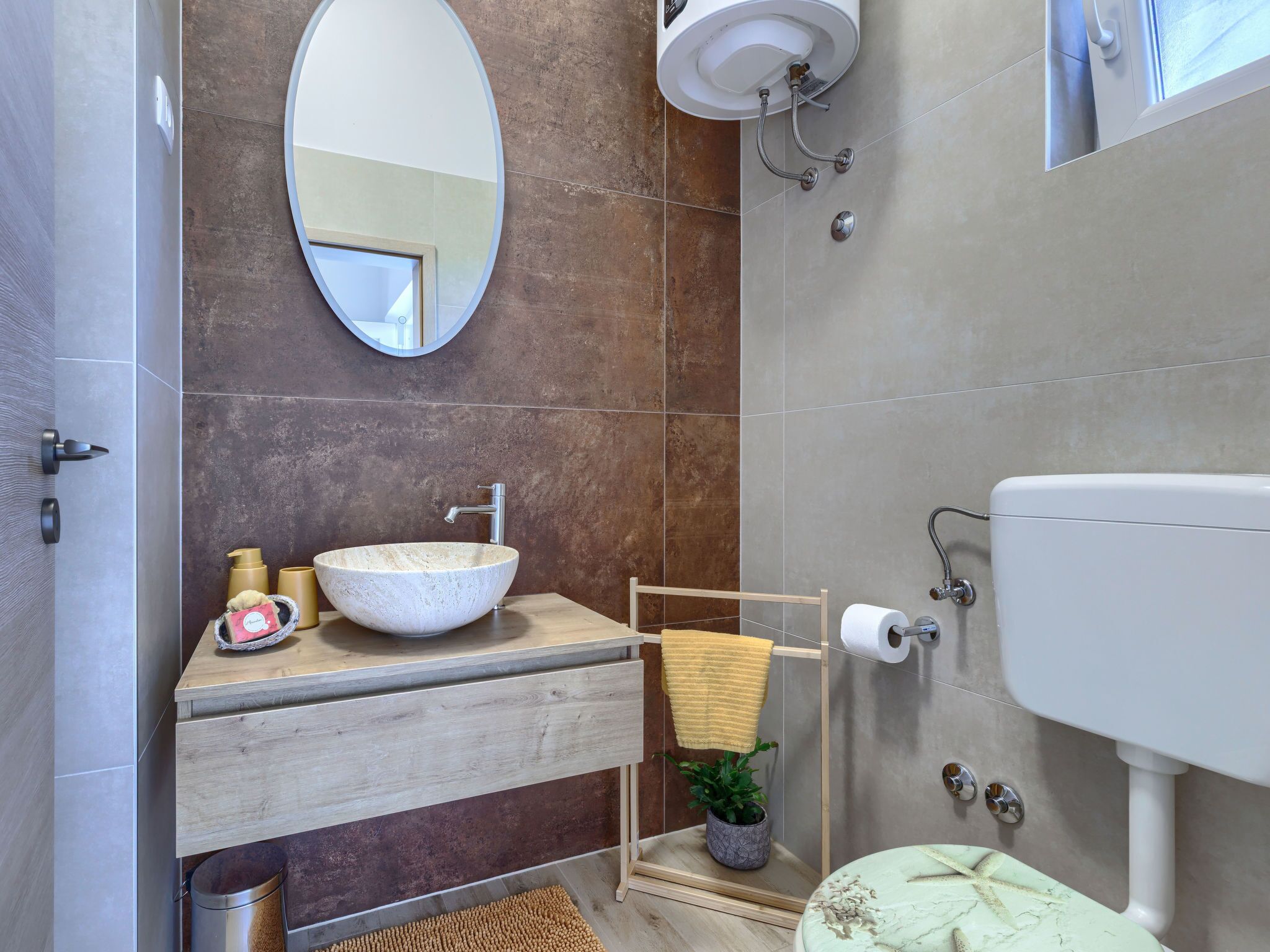A modern bathroom with a large oval mirror, a vessel sink, a toilet, and a shower area.