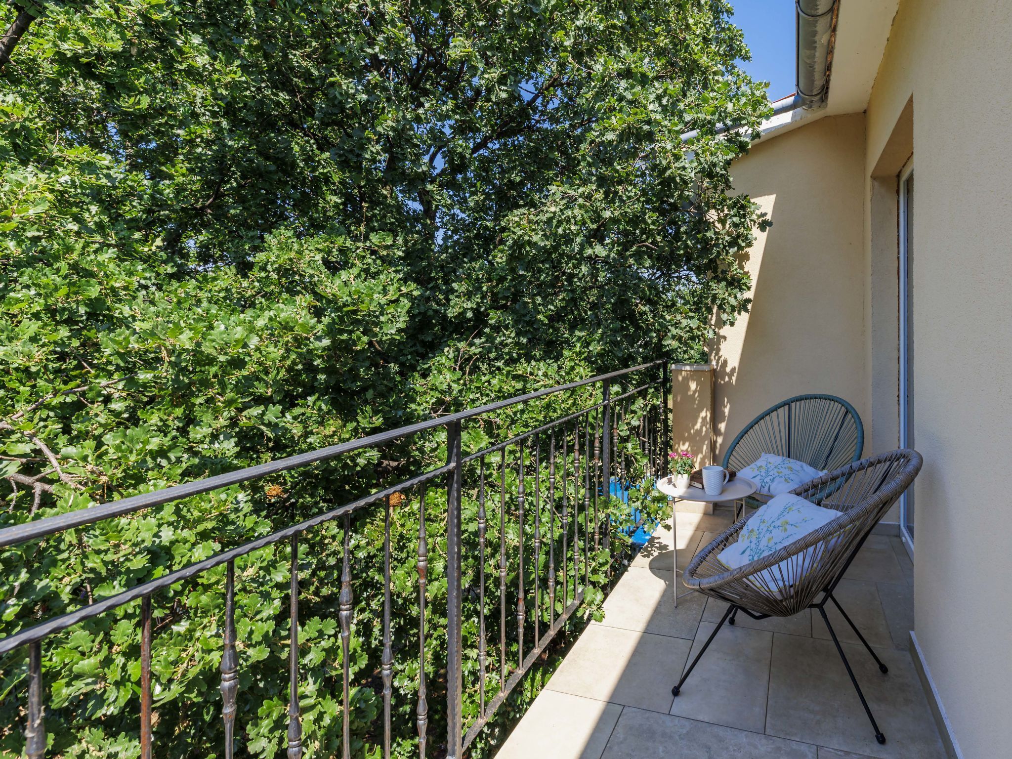 En balkon med gelænder, to fletstole, et lille bord med en plantepotte og en bygning med beige facade.