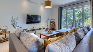 En moderne stue med et fladskærms-tv, en sofa, et sofabord og et stort vindue med udsigt over grønne områder.