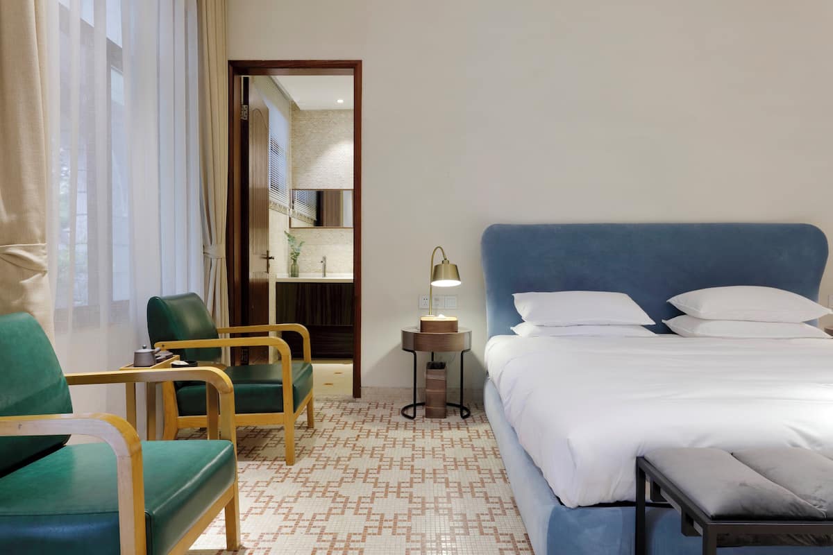 A hotel room with a bed, two green chairs, a small table with a lamp, and a patterned floor.